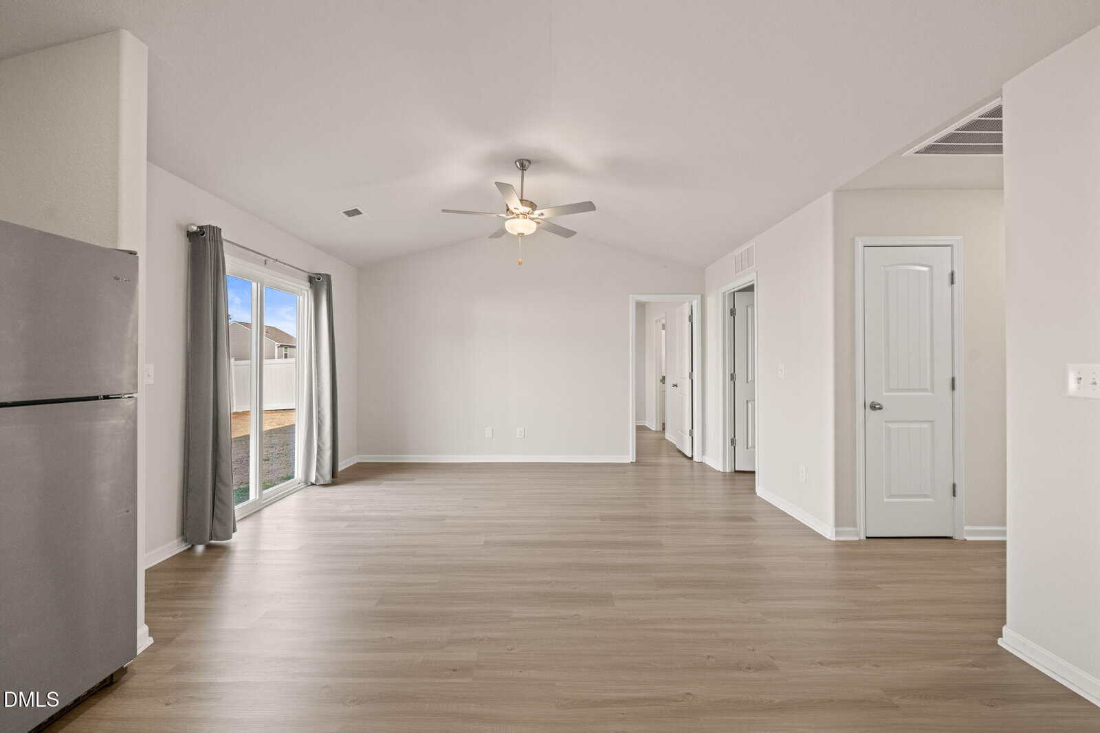 Bright, empty living area with light wood flooring, ceiling fan, sliding glass doors with gray curtains, and several white doors leading to other rooms in a modern home.
