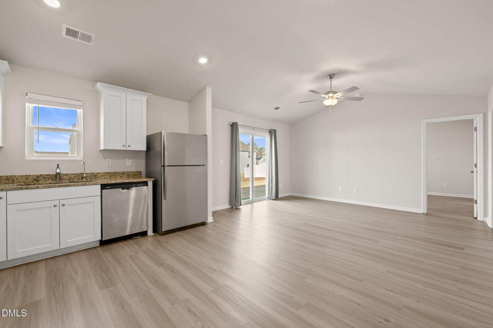 Open-plan kitchen and living area with light wood flooring, stainless steel appliances, white cabinets, a ceiling fan, and sliding glass doors leading to a fenced backyard. A separate room is visible through an open doorway.