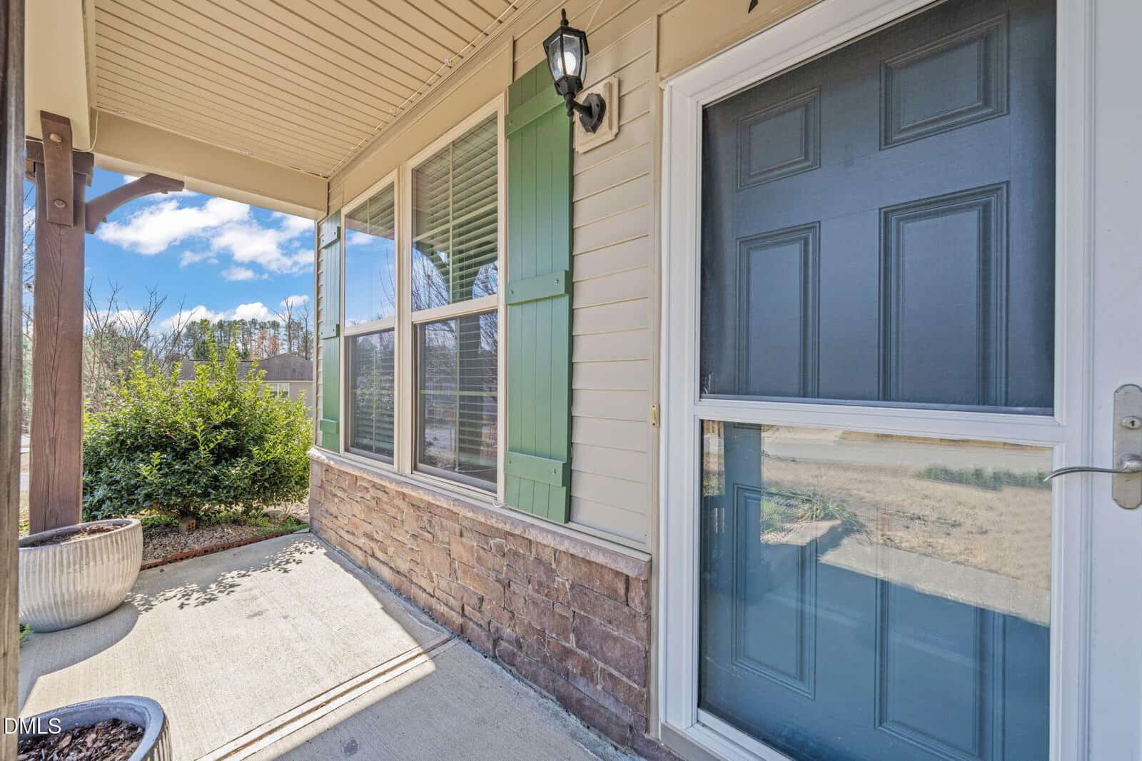 A front porch with a blue door, green shutters, and stone accents. There are potted plants, a lantern-style light fixture, and a concrete walkway. Sunlight casts shadows on the ground.