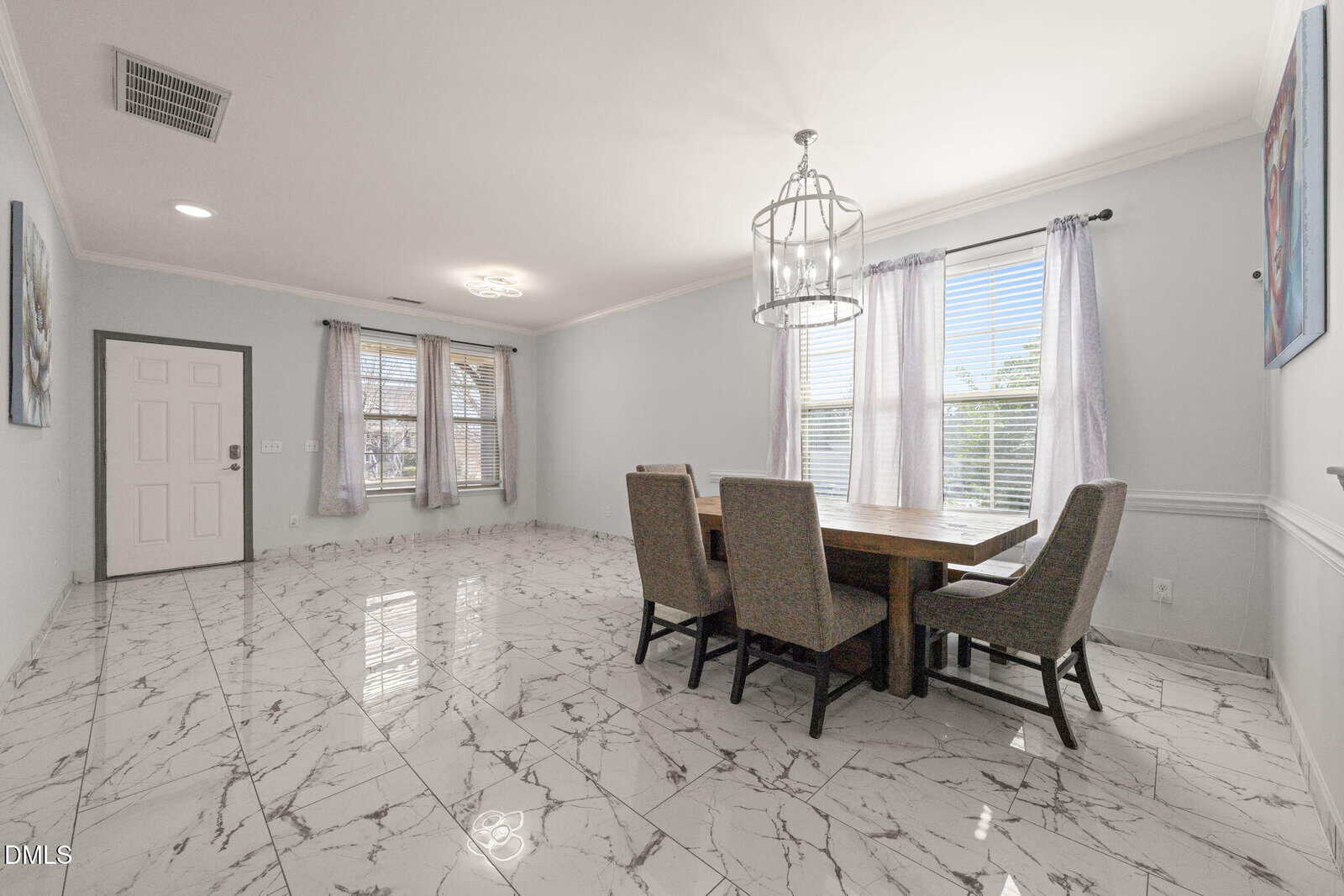 A dining room with a wooden table and six gray upholstered chairs on white marble tile flooring, two large windows with sheer curtains, a modern chandelier, and minimalist decor.