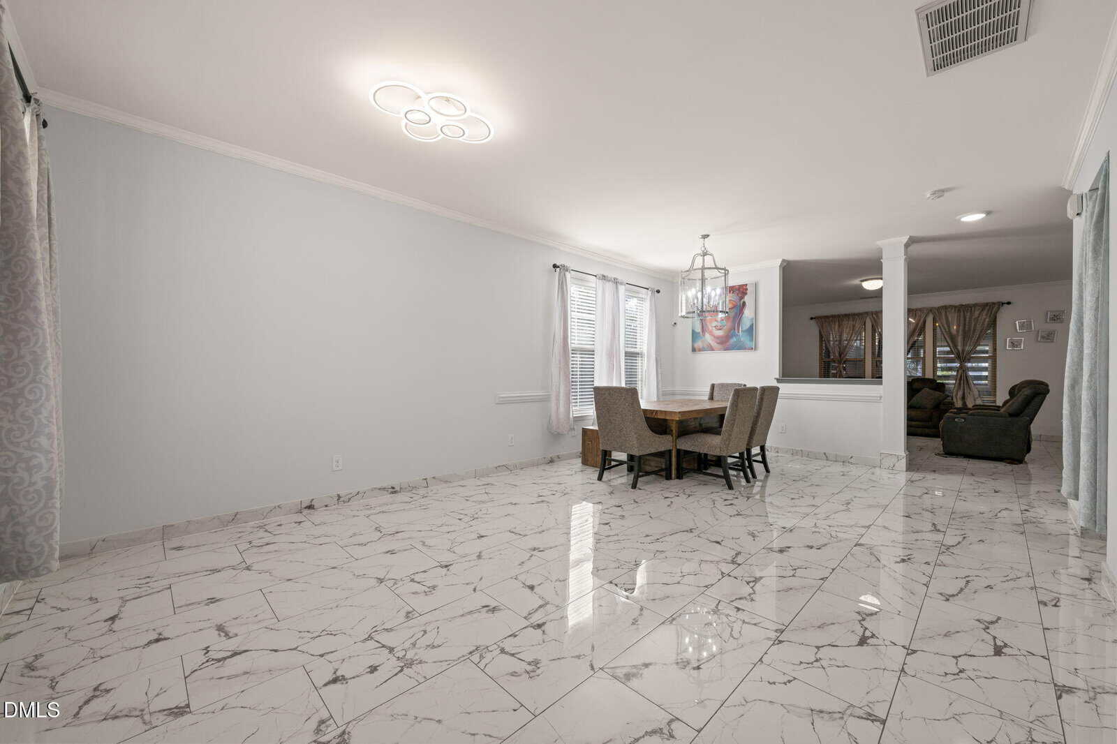 Dining area with a round table and six chairs on a white marble floor, next to large windows with sheer curtains. The space opens to a living room with sofas and a television, separated by two white columns.