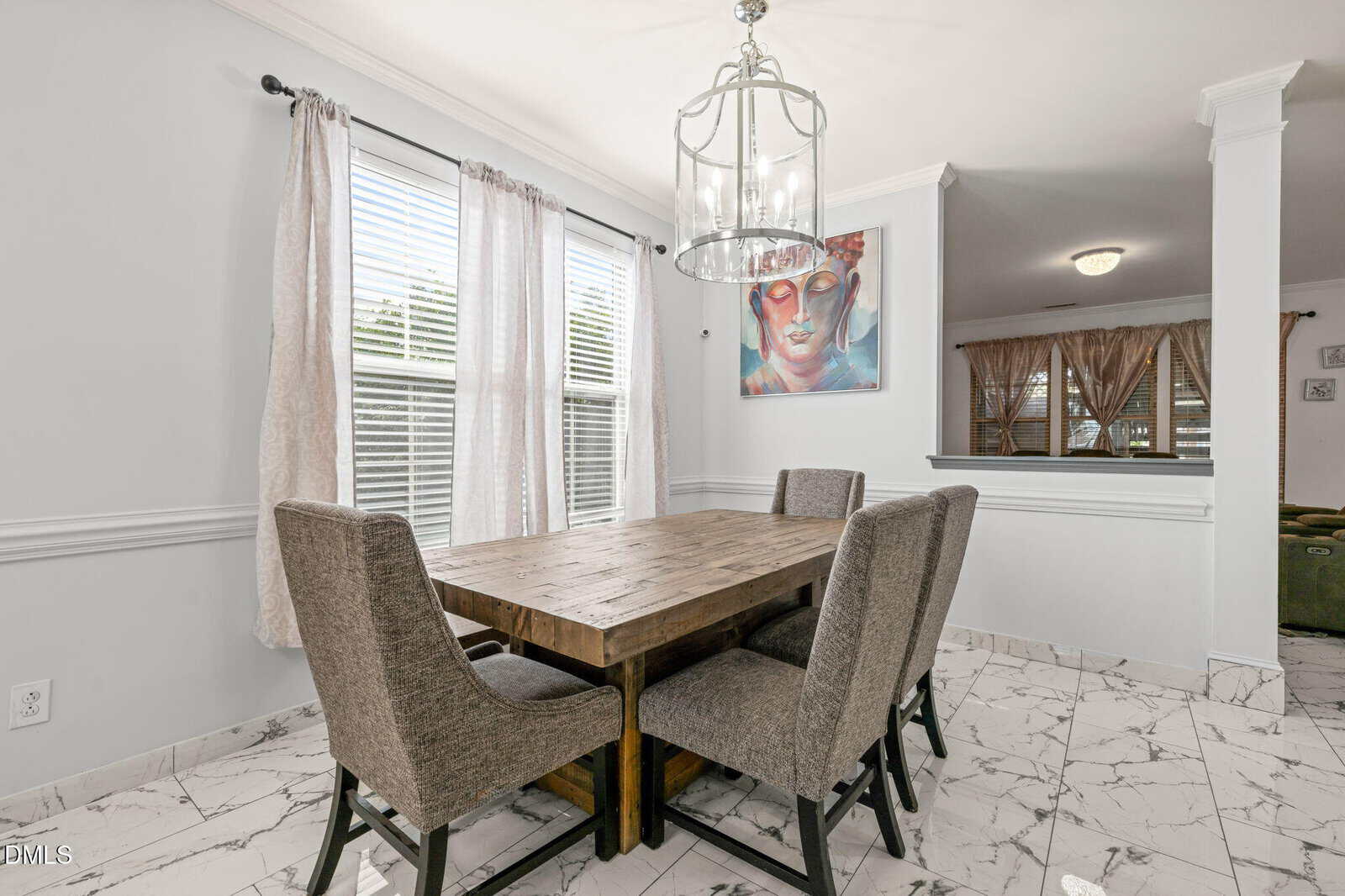 A dining area with a wooden table, four upholstered chairs, marble tile floor, large windows with curtains, and a modern chandelier. A colorful painting hangs on the wall beside the table.