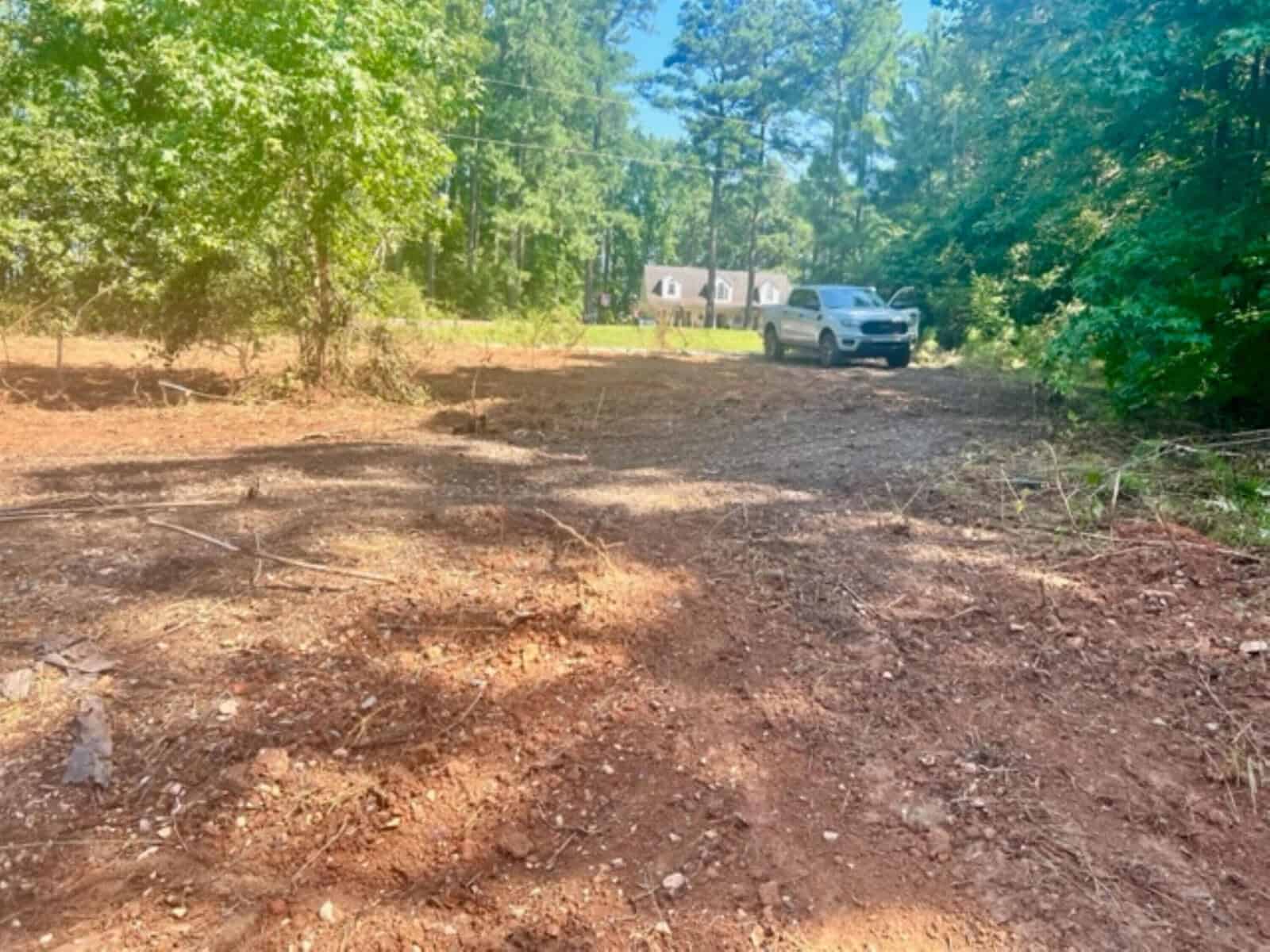 A cleared patch of dirt and scattered debris is surrounded by trees. A white SUV is parked on the right side, and a house on a large grassy lot is visible in the background. The sky is clear and sunny.