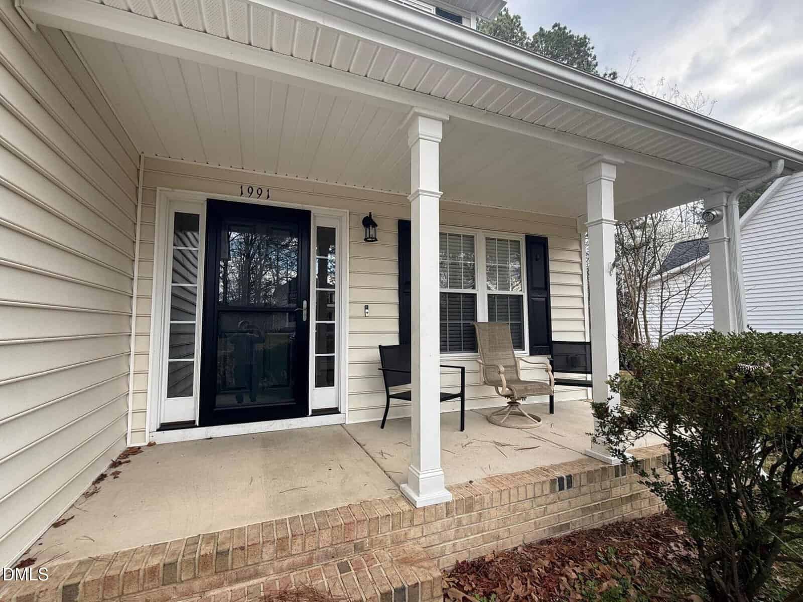 A small covered front porch with white pillars, beige siding, a black storm door, two chairs, a small table, house number 1991, and neatly trimmed bushes along the brick steps.