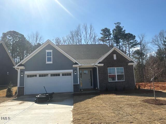 A single-story house at 230 Sawtooth Oak Lane in Youngsville features gray siding, brick accents, and a two-car garage, sitting on a lawn with sparse grass. A black hose rests on the driveway, while leafless trees stand in the background under a clear sky.