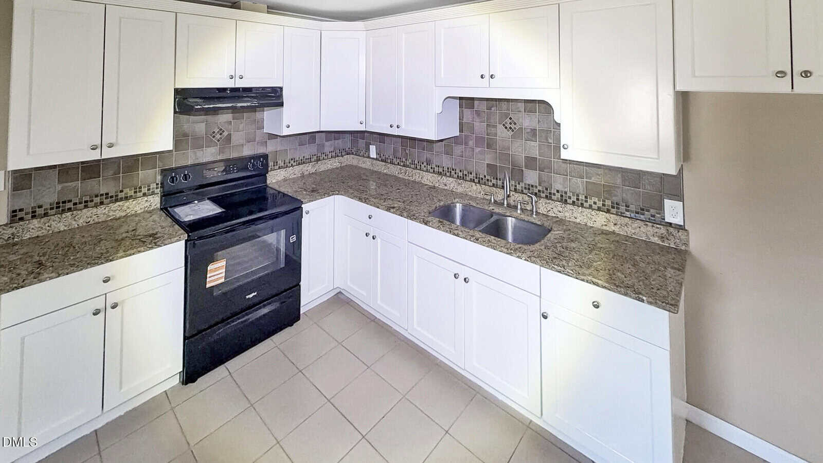 A kitchen with white cabinets, a black stove and oven, a double sink, granite countertops, and beige tile flooring. The backsplash features a pattern of neutral-colored tiles.