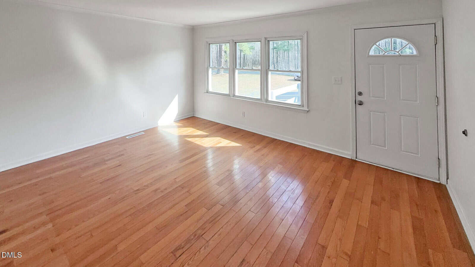 Bright, empty room with light wood floors, three windows letting in sunlight, and a white front door with a small window. Walls are plain white and there are no furnishings or decorations.