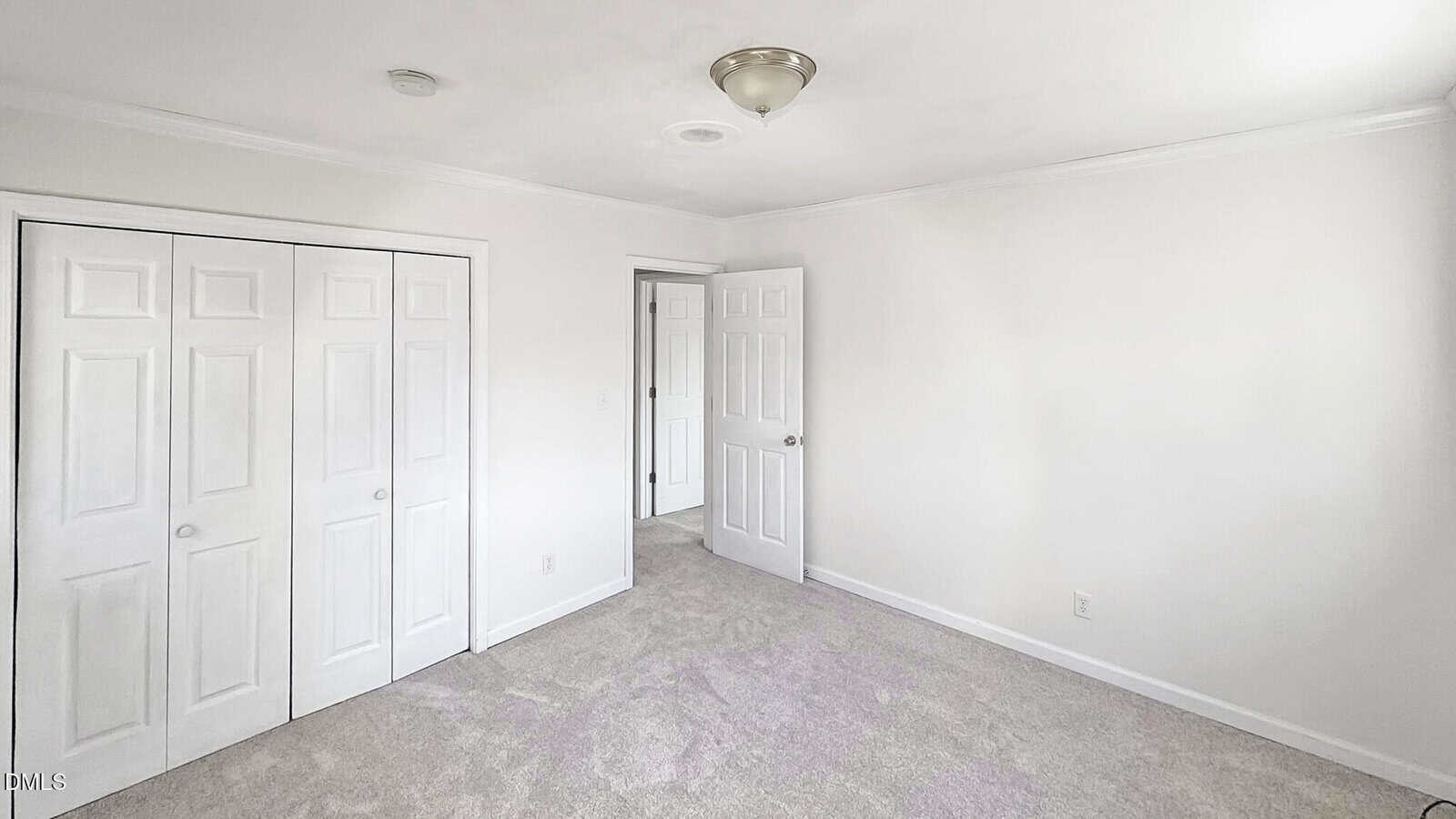 Empty bedroom with white walls, beige carpet, a ceiling light fixture, closed double closet doors, and an open door leading to a hallway. Natural light is coming in from the right side.
