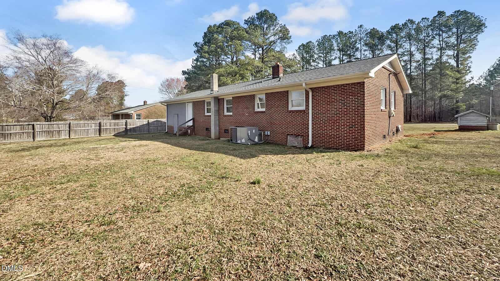 A brick house with a fenced backyard, HVAC unit, and a grassy lawn, surrounded by trees under a partly cloudy sky.