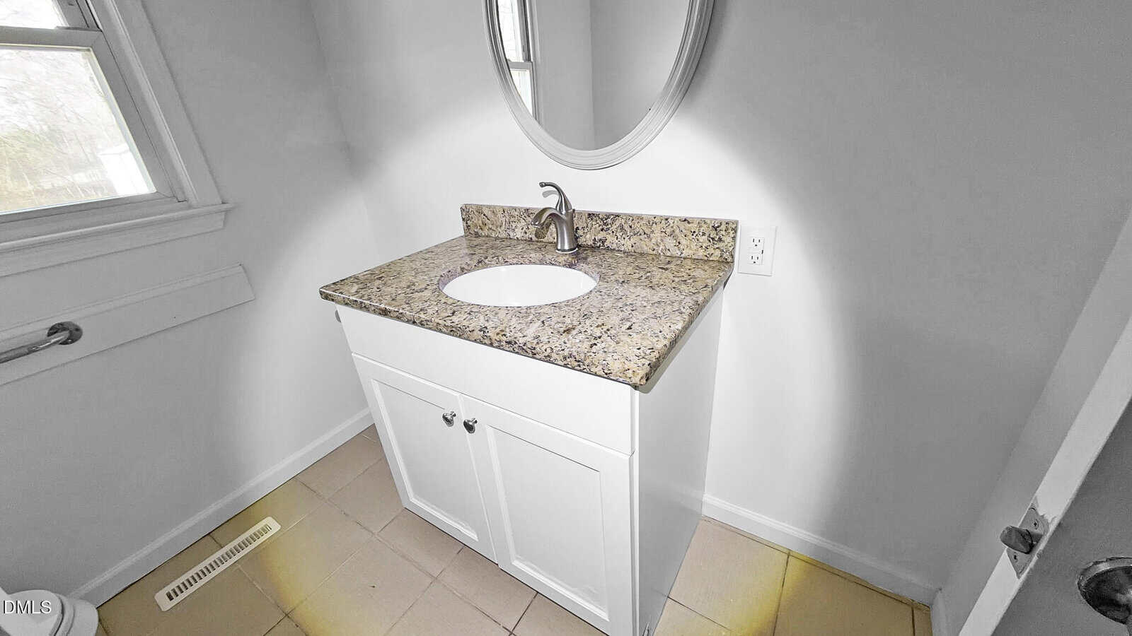 A bathroom with a white vanity, granite countertop, oval sink, silver faucet, round mirror, tan tile floor, and a window on the left.