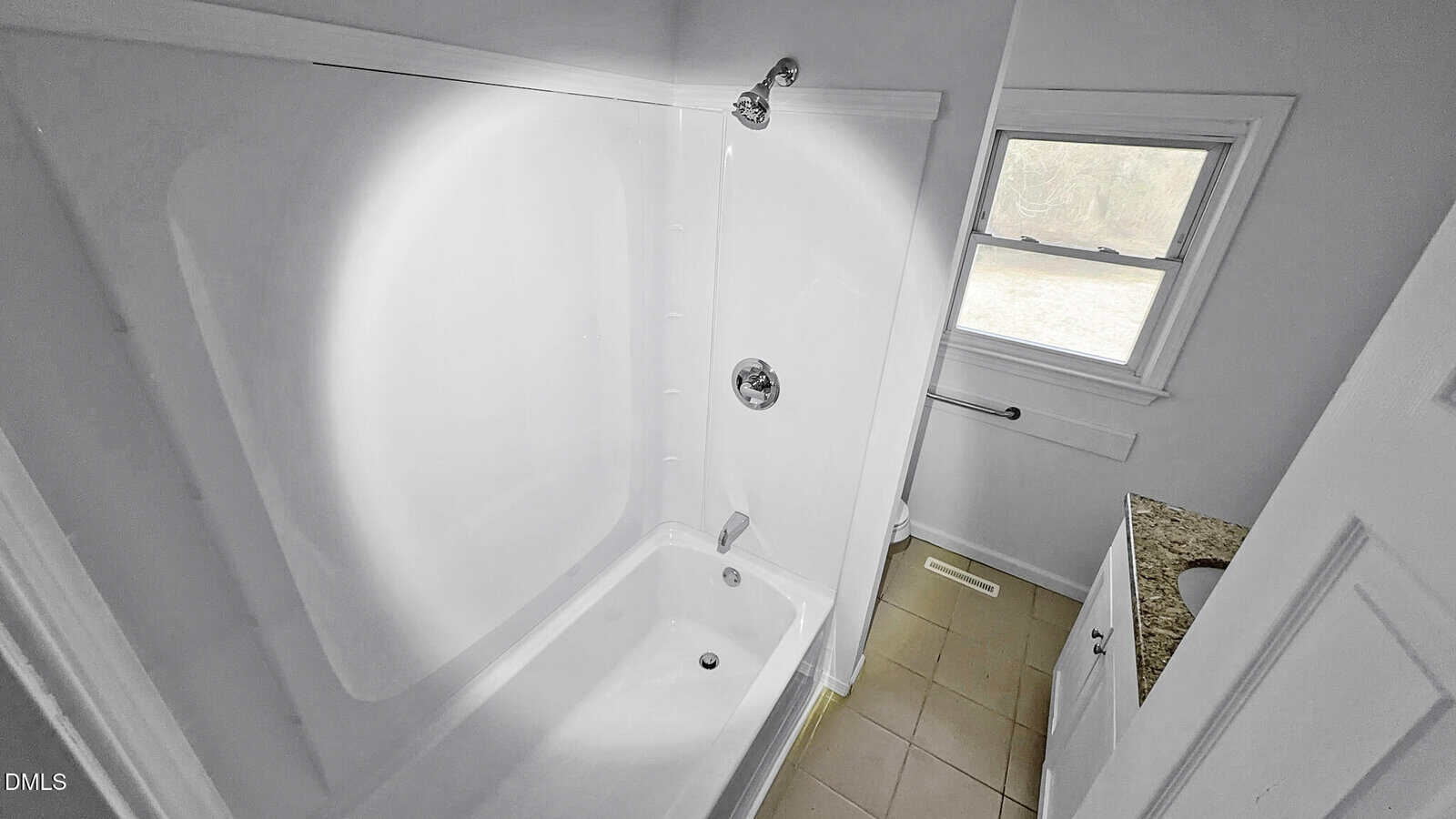 A bathroom with a white bathtub and shower, a window, a small sink with a granite countertop, a toilet, and beige tile flooring. The walls are white and the area appears clean and minimalistic.
