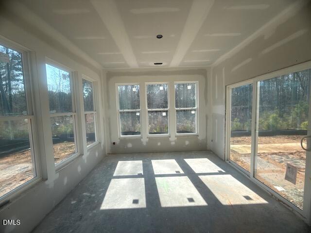A room under construction with unfinished drywall, large windows on three walls, and sunlight casting shadows on the concrete floor. Trees and dirt are visible outside.