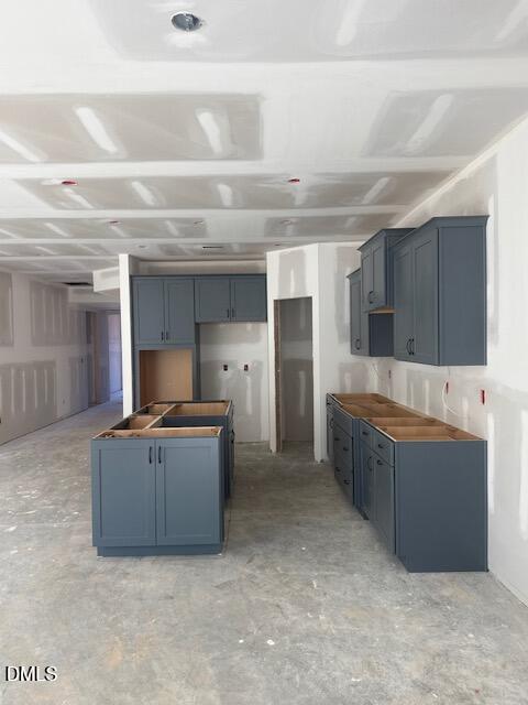 Unfinished kitchen with gray cabinets installed and no countertops. The walls and ceiling are covered in unpainted drywall with visible seams. The floor is bare concrete, and there are no appliances or fixtures installed yet.