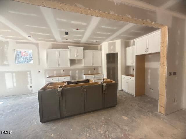 Unfinished kitchen with drywall, unpainted walls, white cabinets, a gray center island, and no appliances or countertops installed. Construction materials and dust are visible on the floor.
