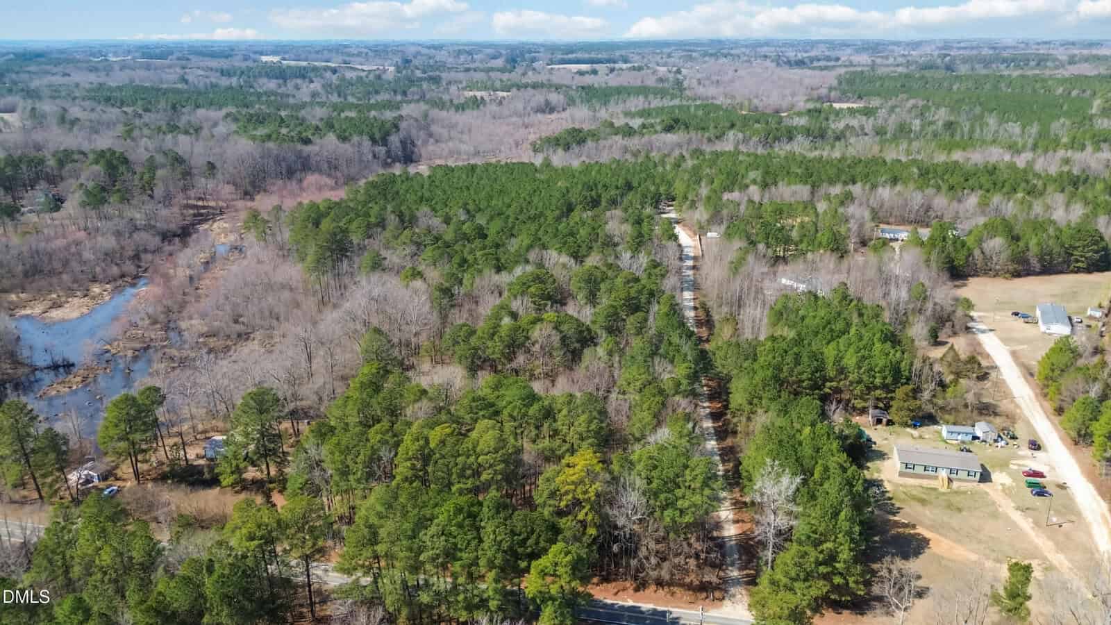 Aerial view of a rural area with dense forests, a river or creek on the left, and scattered houses along dirt roads. The landscape is mostly green with some leafless trees, and open fields are visible near the homes.