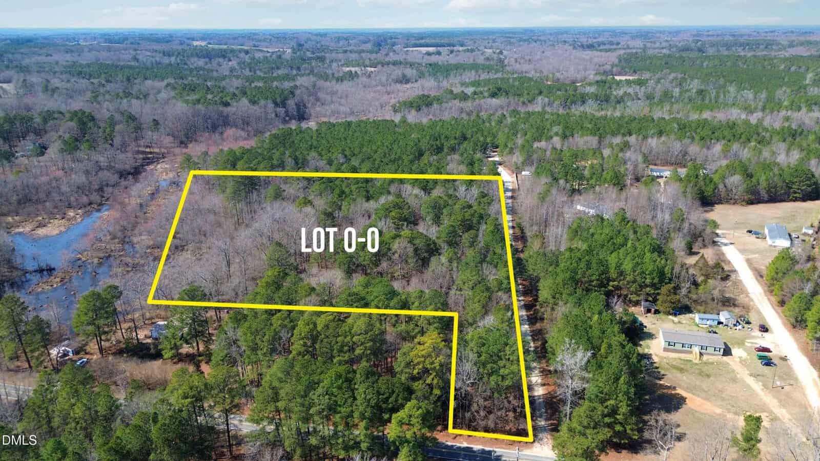 Aerial view of a wooded lot outlined in yellow, labeled “LOT 0-0,” located off 0 Old Halifax Road near Zebulon. The property is surrounded by dense trees, with a lake on the left and a few houses, open land, and dirt roads nearby. Sky is partly cloudy.