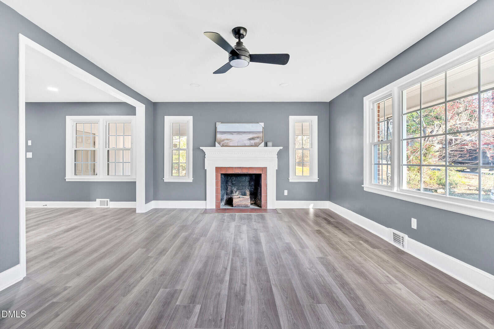 A bright living room with gray walls, large windows, and wood-look flooring features a white fireplace with a black ceiling fan centered above. An adjacent room is visible through a wide entryway.