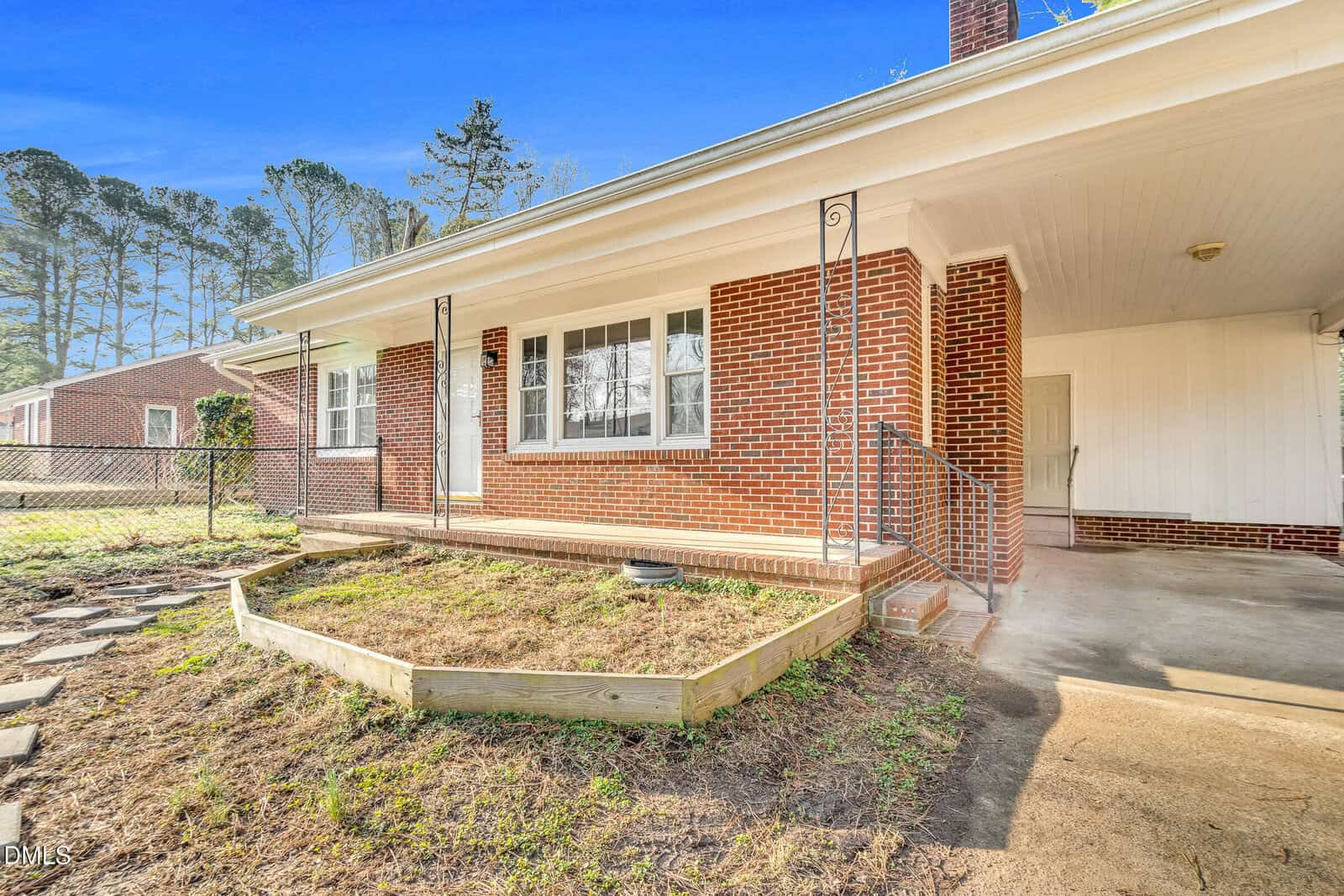 Single-story brick house with a covered front porch, white trim, three windows, metal railings, and a small raised garden bed. The yard has some grass and a stone path, and there are trees in the background.