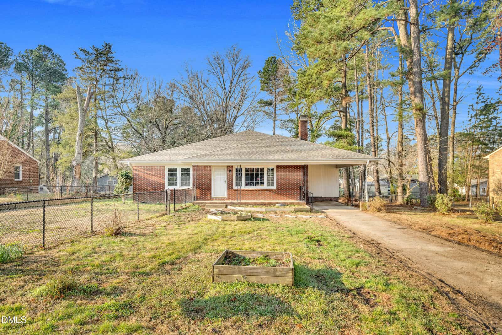 Single-story red brick house with a gray roof, front porch, and white trim, set among tall trees on 1249 Oakridge Avenue in Henderson. A driveway and chain-link fence run along the front yard, which has a small raised garden bed. The sky is clear and blue.