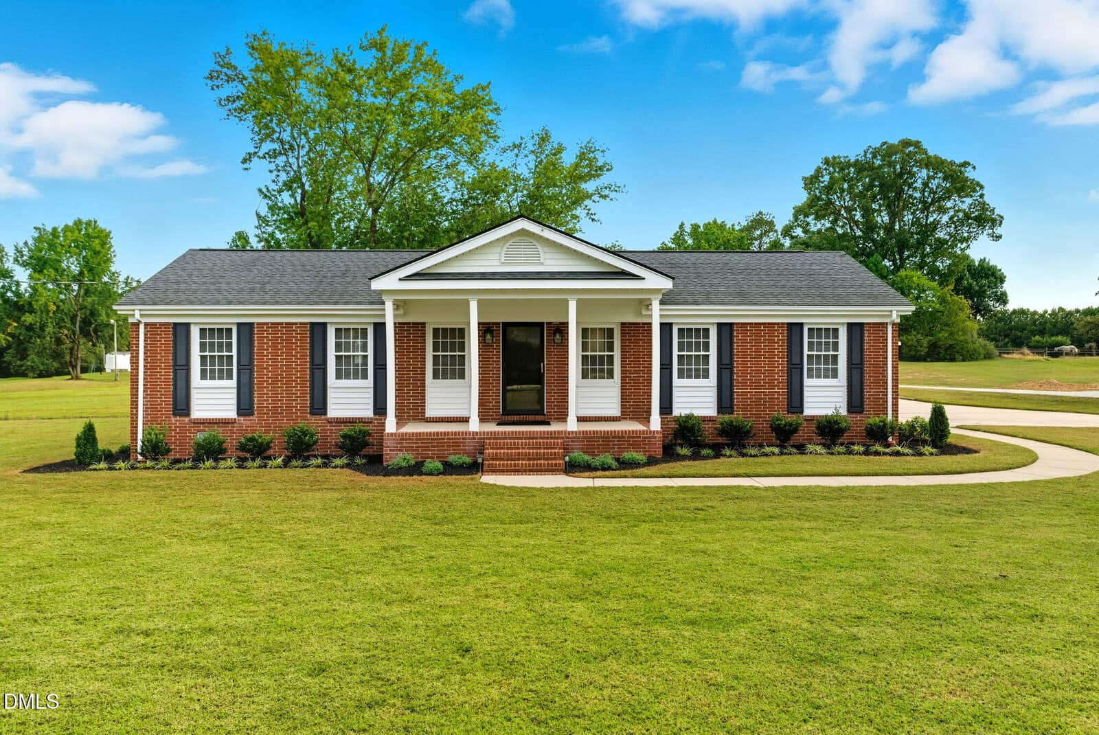 Single-story brick house with white trim, six front windows, a covered entryway with columns, and a front lawn. Located at 2238 Tarboro Road in Youngsville, this home features a curved concrete walkway leading to the steps, while trees are visible in the background under a blue sky.