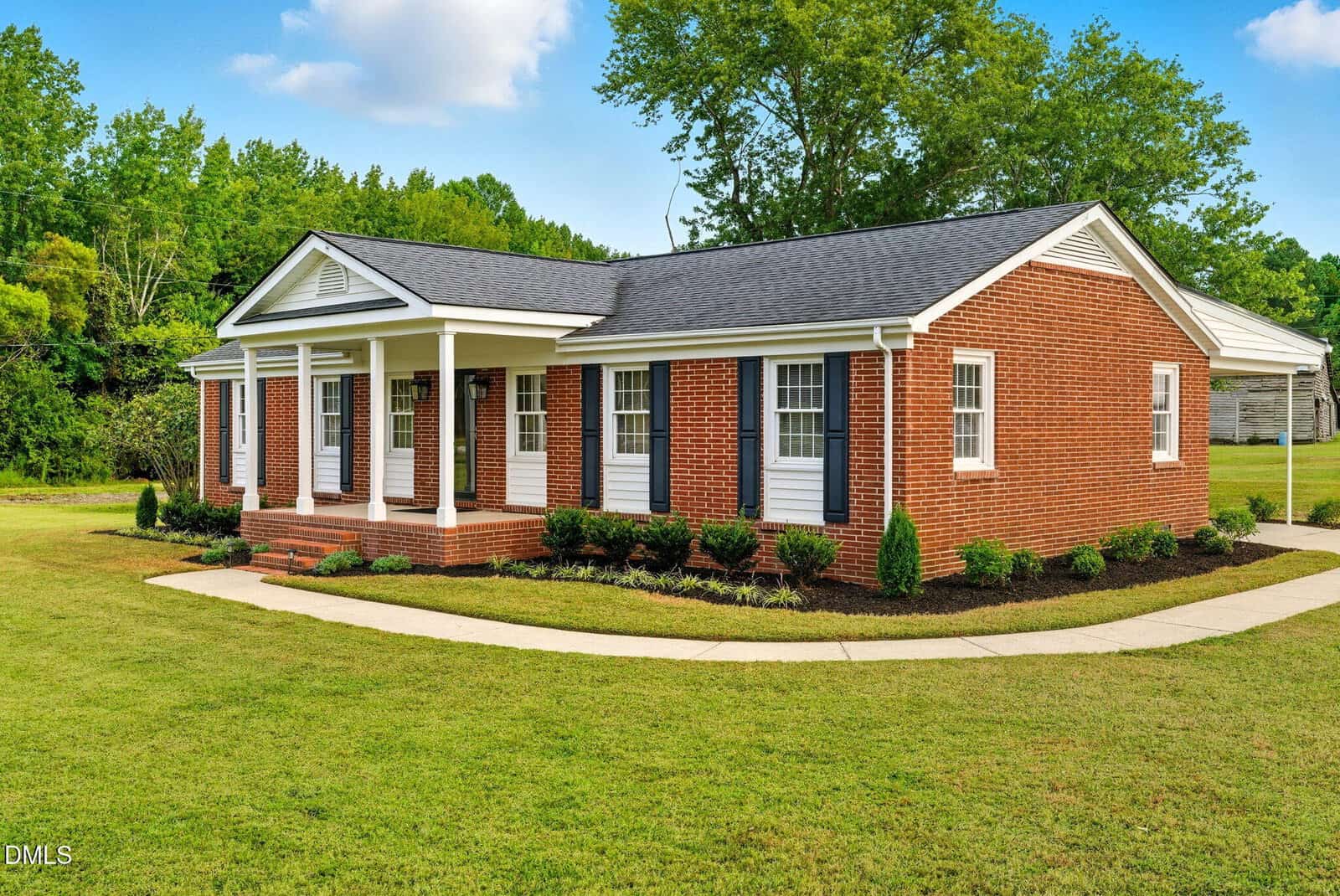 A single-story red brick house with white trim, black shutters, and a covered front porch. The house sits on a well-maintained lawn with a sidewalk and has small shrubs planted along the front.