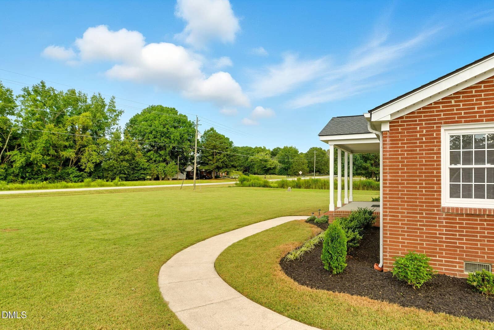 A curved concrete walkway leads to a brick house with white trim, bordered by bushes and mulch. The large front lawn is bordered by trees under a blue sky with scattered clouds.