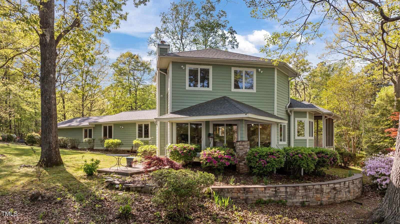 A two-story house at 373 Sagamore Drive in Louisburg features light green siding and white trim, surrounded by trees and landscaped bushes on a grassy yard. A stone retaining wall and patio furniture enhance the inviting garden setting.
