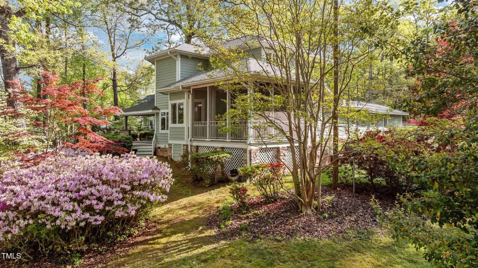 A charming light green house with a wraparound porch sits among blooming azaleas and lush trees in a well-landscaped garden under a bright sky.