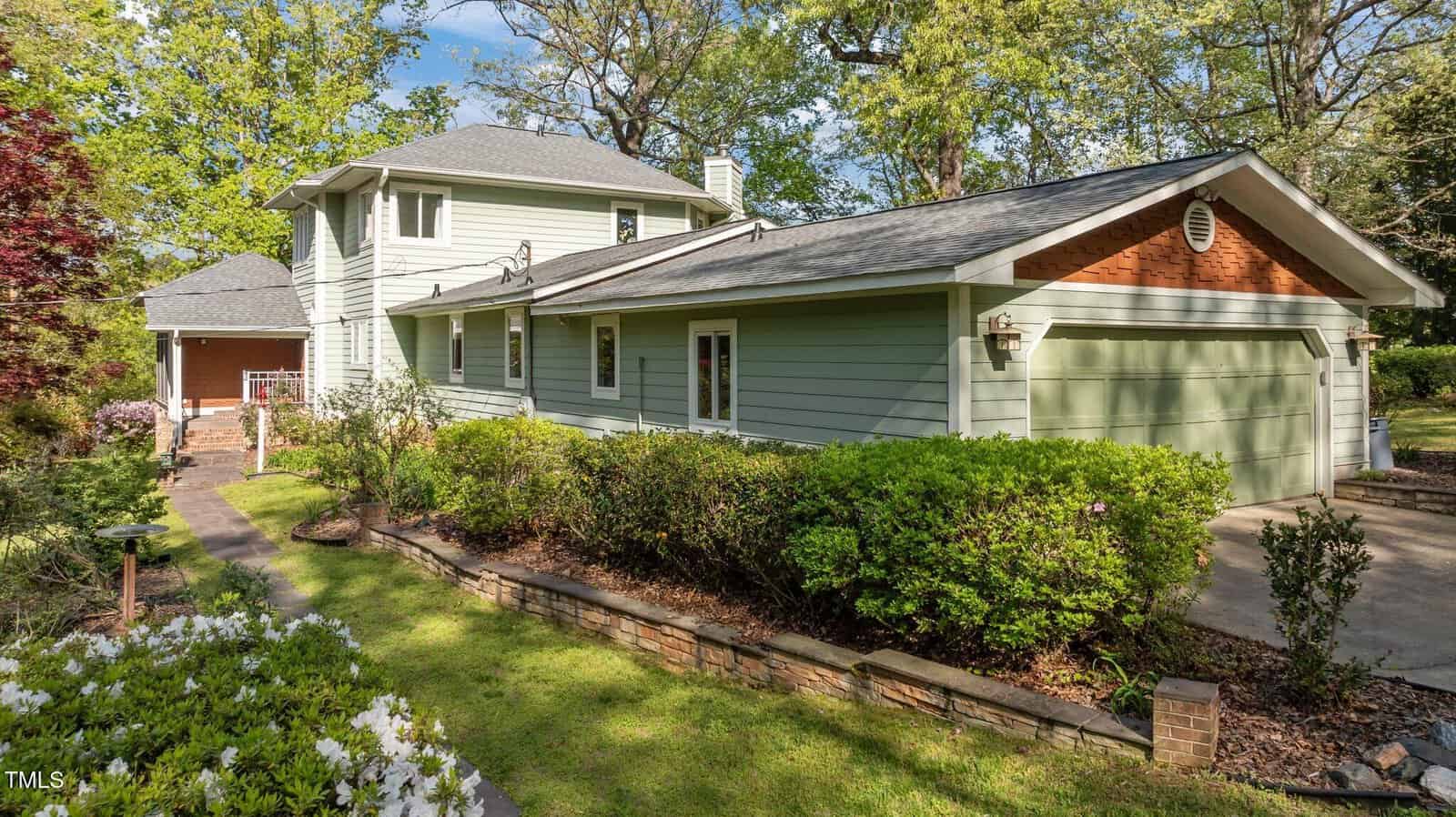 A two-story house at 373 Sagamore Drive in Louisburg with light green siding, white trim, and a gray roof, featuring a double garage and surrounded by landscaped gardens, trees, and a pathway leading to the entrance.