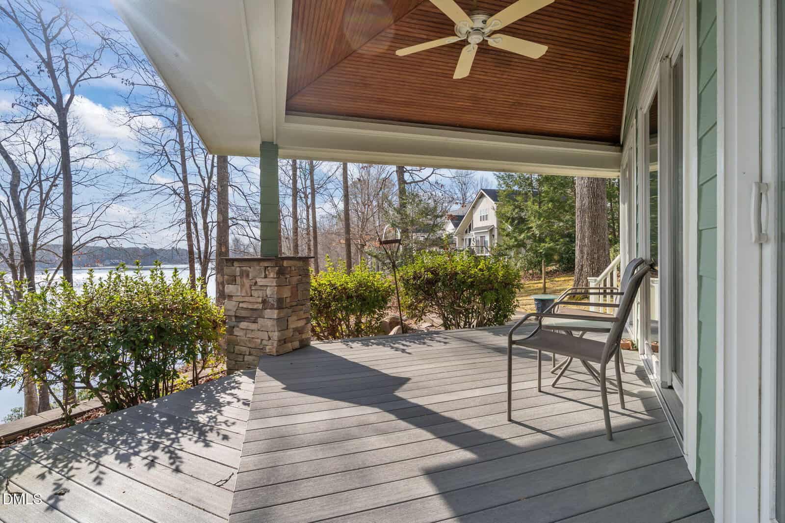 A cozy porch with a ceiling fan, two chairs, and a view of trees, a lake, and nearby houses. Bushes line the edge of the porch, and sunlight filters through the branches.