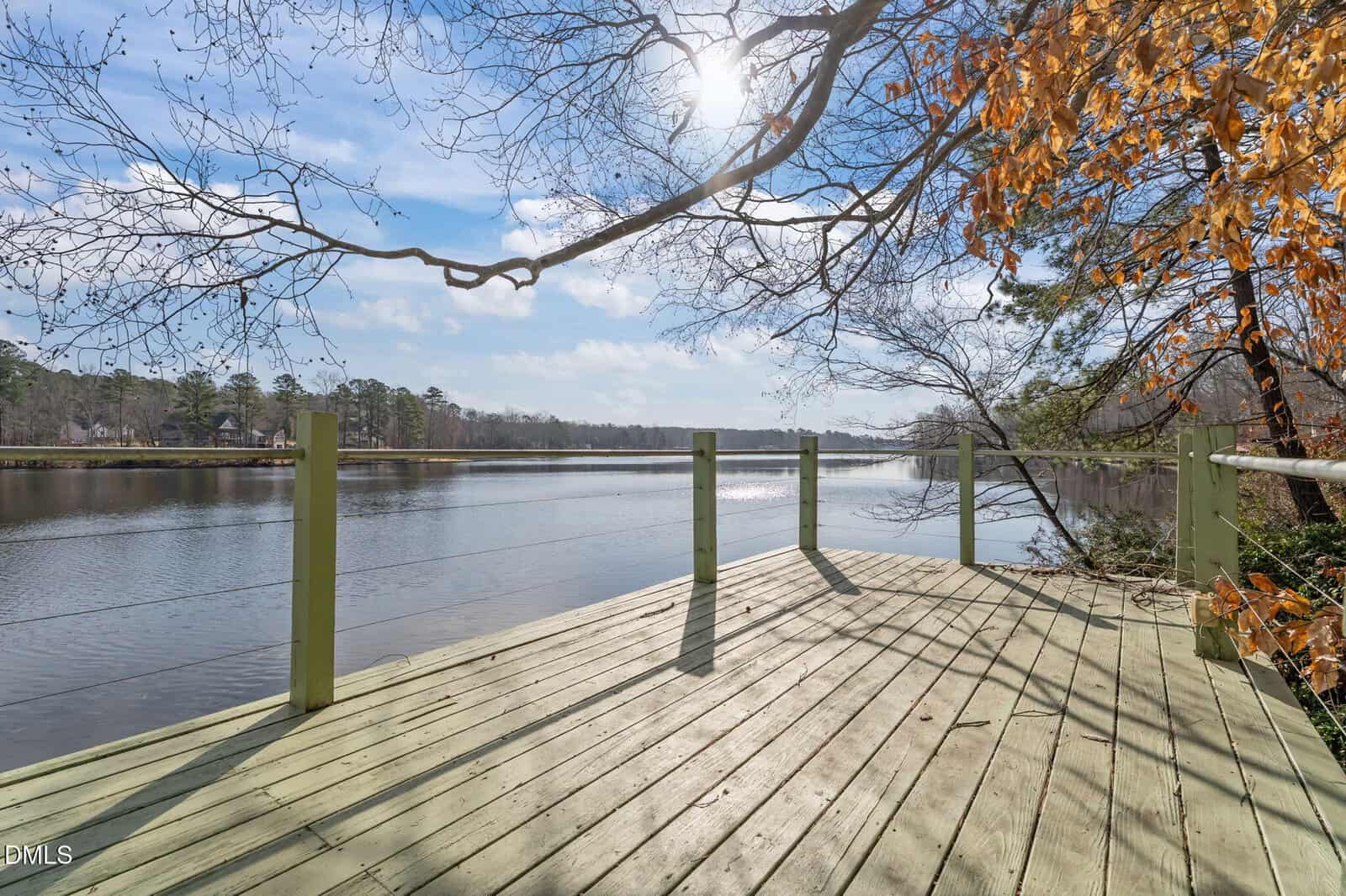 A wooden deck overlooking a calm lake, surrounded by trees with some autumn leaves. The sun is shining through branches, and the sky is partly cloudy.
