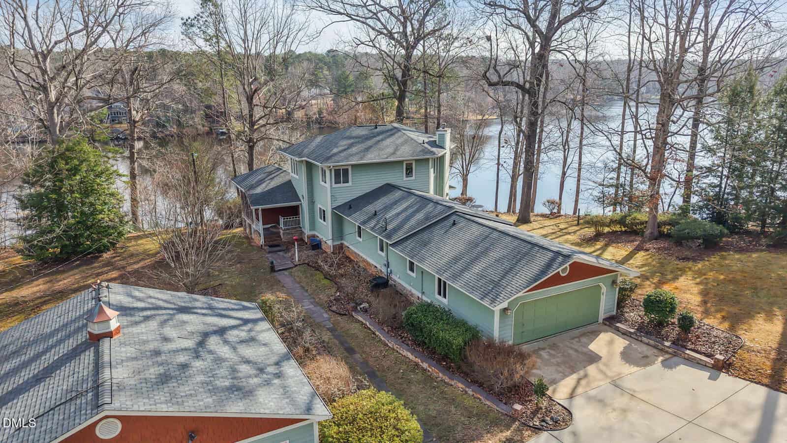 A two-story green house with a gray roof sits beside a lake, surrounded by leafless trees and a manicured yard. A driveway leads to an attached double garage, and a detached structure is visible in the foreground.