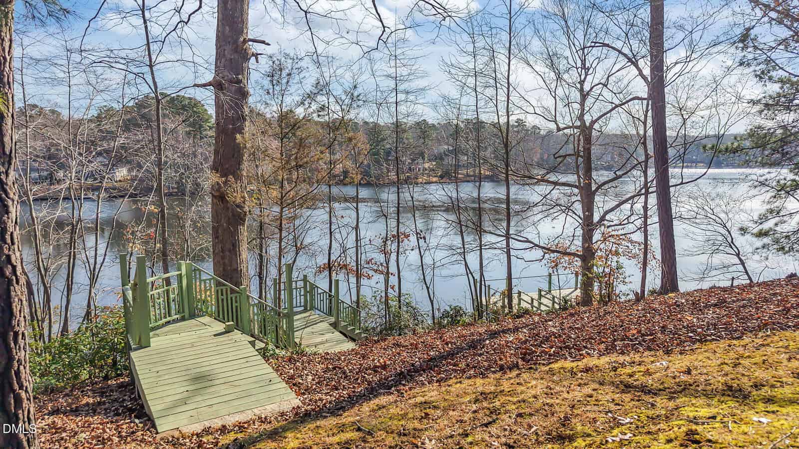 A wooden staircase with green railings leads down a leaf-covered slope to a calm lake, surrounded by bare trees and forested shoreline under a partly cloudy sky.