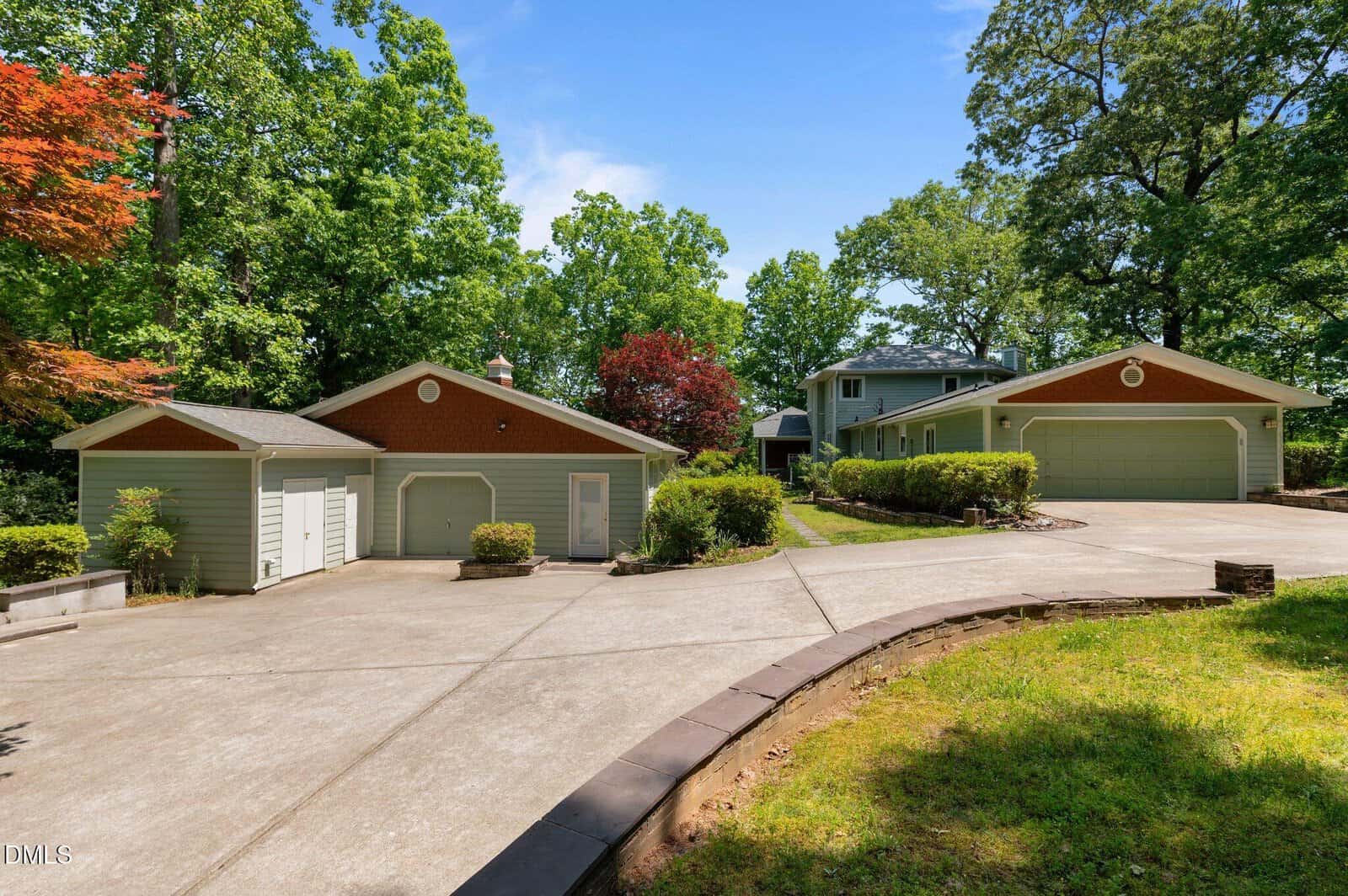 A wide concrete driveway at 373 Sagamore Drive in Louisburg leads to two separate green garages with white doors, surrounded by trees and shrubs, with a two-story house visible in the background under a clear, blue sky.