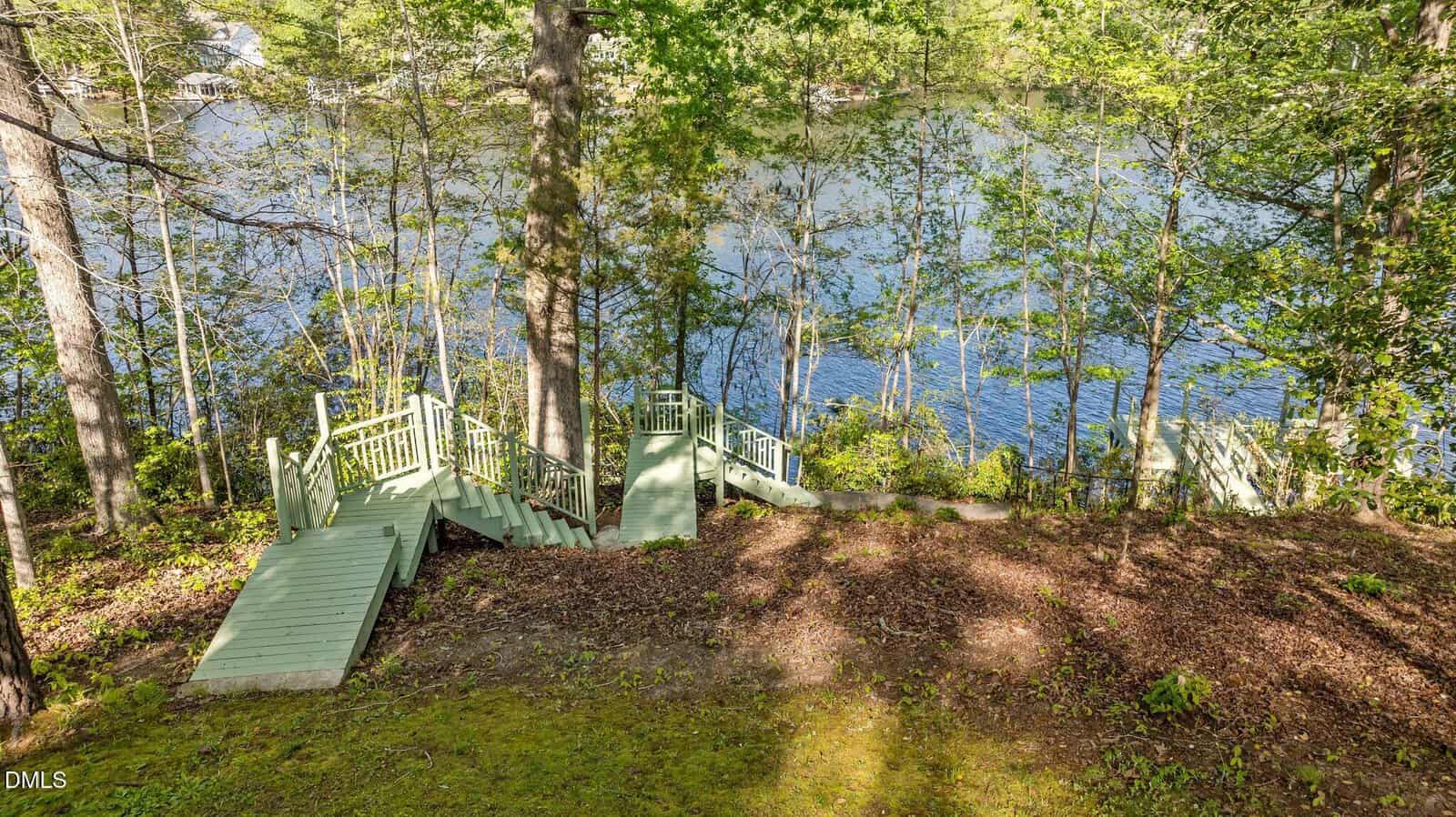 Wooden stairs painted light green descend a forested slope toward a calm lake, surrounded by lush trees and dappled sunlight.