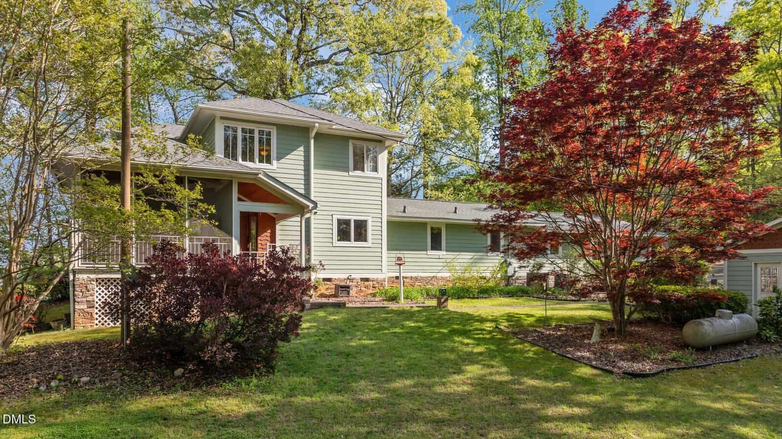 Two-story light green house with a covered front porch, surrounded by trees and landscaped garden, including a red-leafed tree and green lawn, under a clear blue sky.