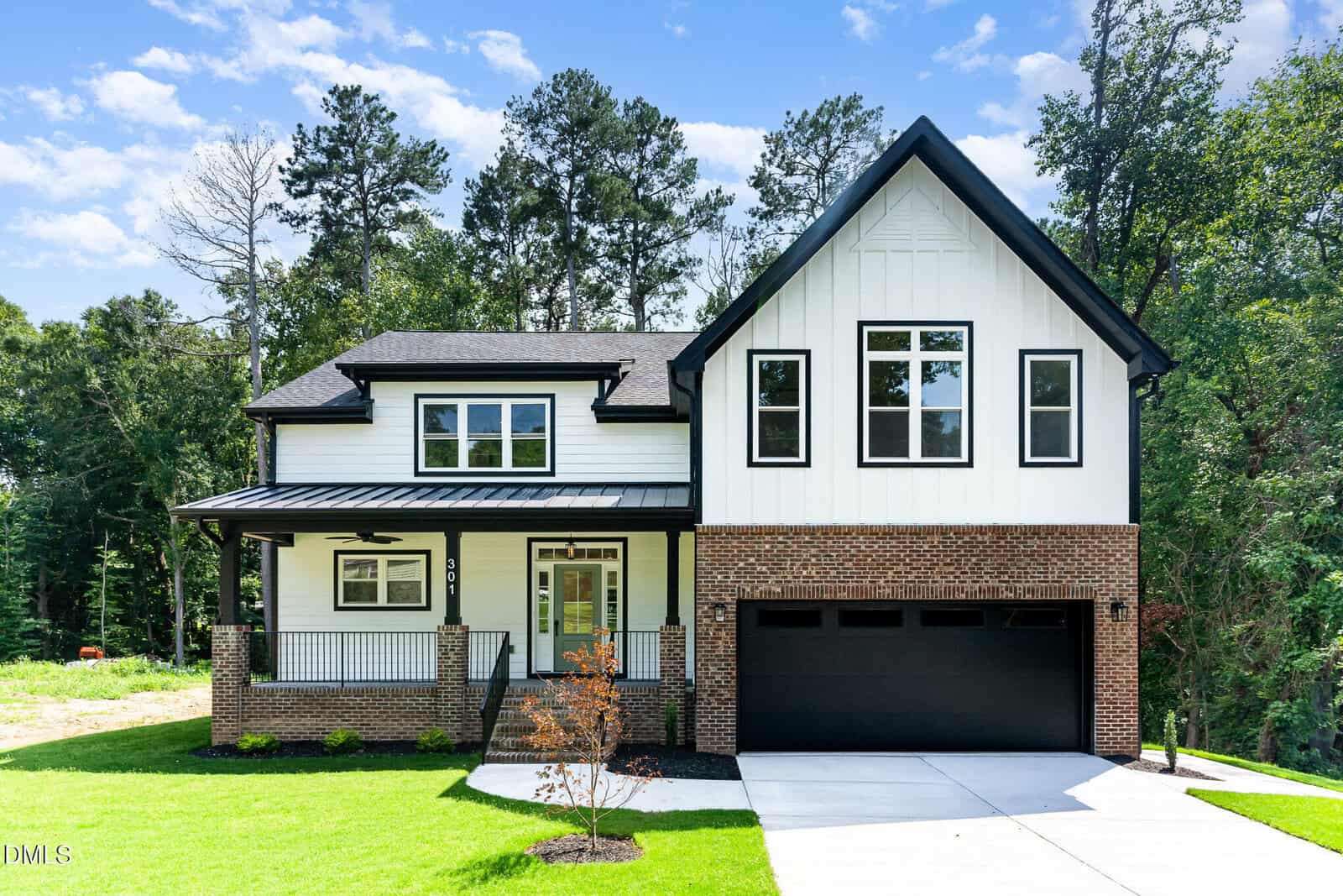 Modern two-story house with a covered front porch, brick and white siding exterior, black trim, large windows, and a two-car garage, set against a backdrop of trees on a sunny day.