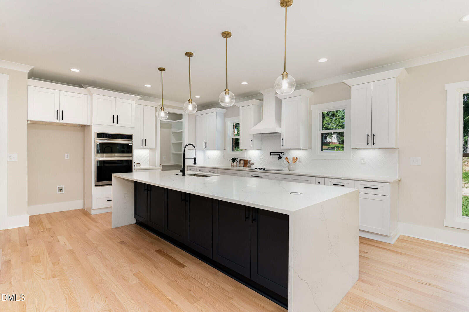 Modern kitchen with white cabinets, black island, marble countertops, pendant lights, hardwood floors, stainless steel appliances, and large windows letting in natural light.