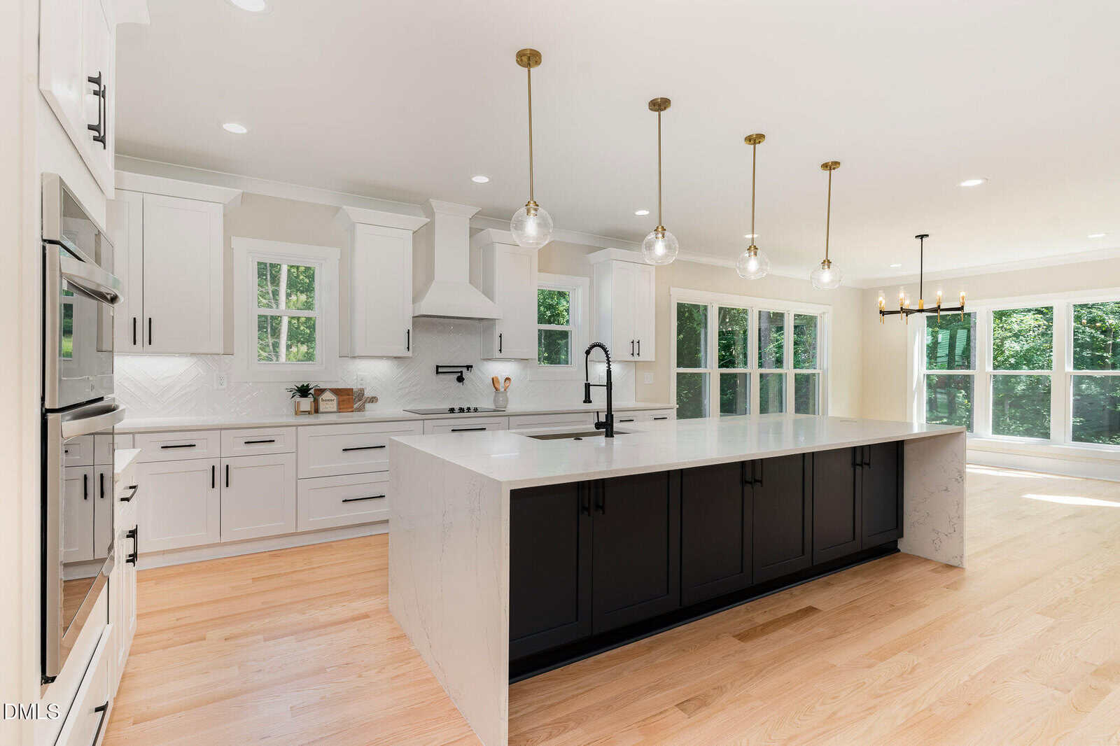 Modern kitchen with white cabinets, black island, quartz countertops, stainless steel appliances, wood flooring, pendant lights above the island, and large windows providing natural light in an open, bright space.