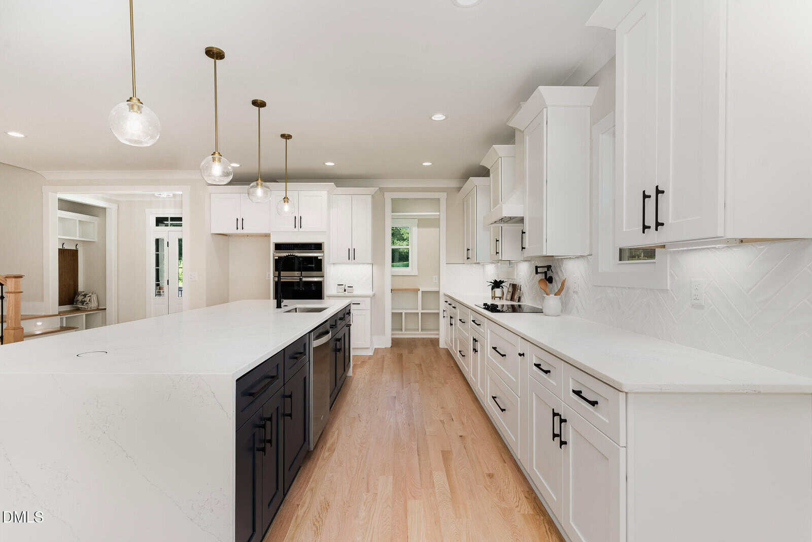 Modern kitchen with white cabinets, black island, light wood flooring, pendant lights, stainless steel appliances, and white countertops. There are windows, open shelves, and a small built-in desk area in the background.