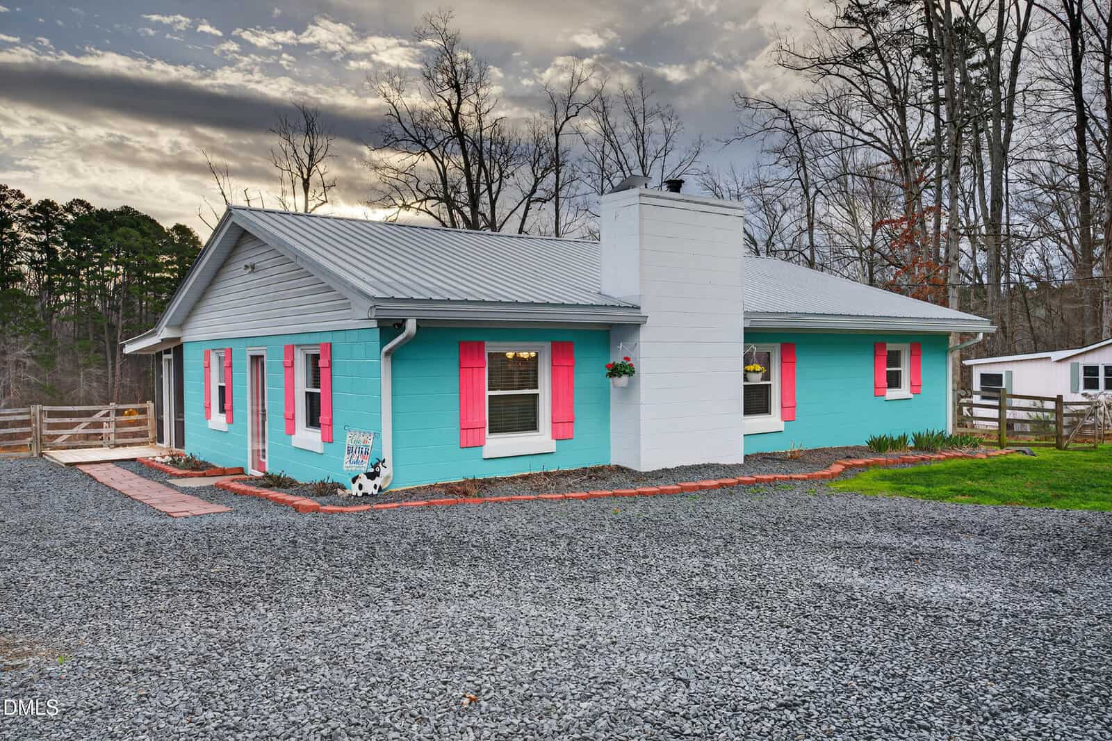 A single-story house with turquoise exterior walls, white trim, and pink shutters. There is a white chimney, gravel driveway, and a small fenced yard with trees in the background under a cloudy sky.