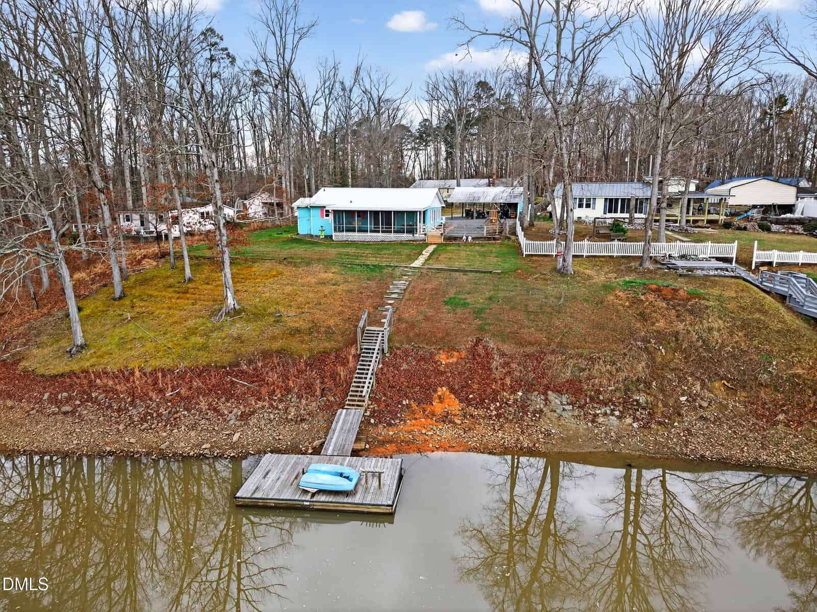 Aerial view of a lakeside property with a green house, wooden dock extending into the water, bare trees, and several neighboring homes. The lawn is patchy with reddish soil visible near the shoreline.
