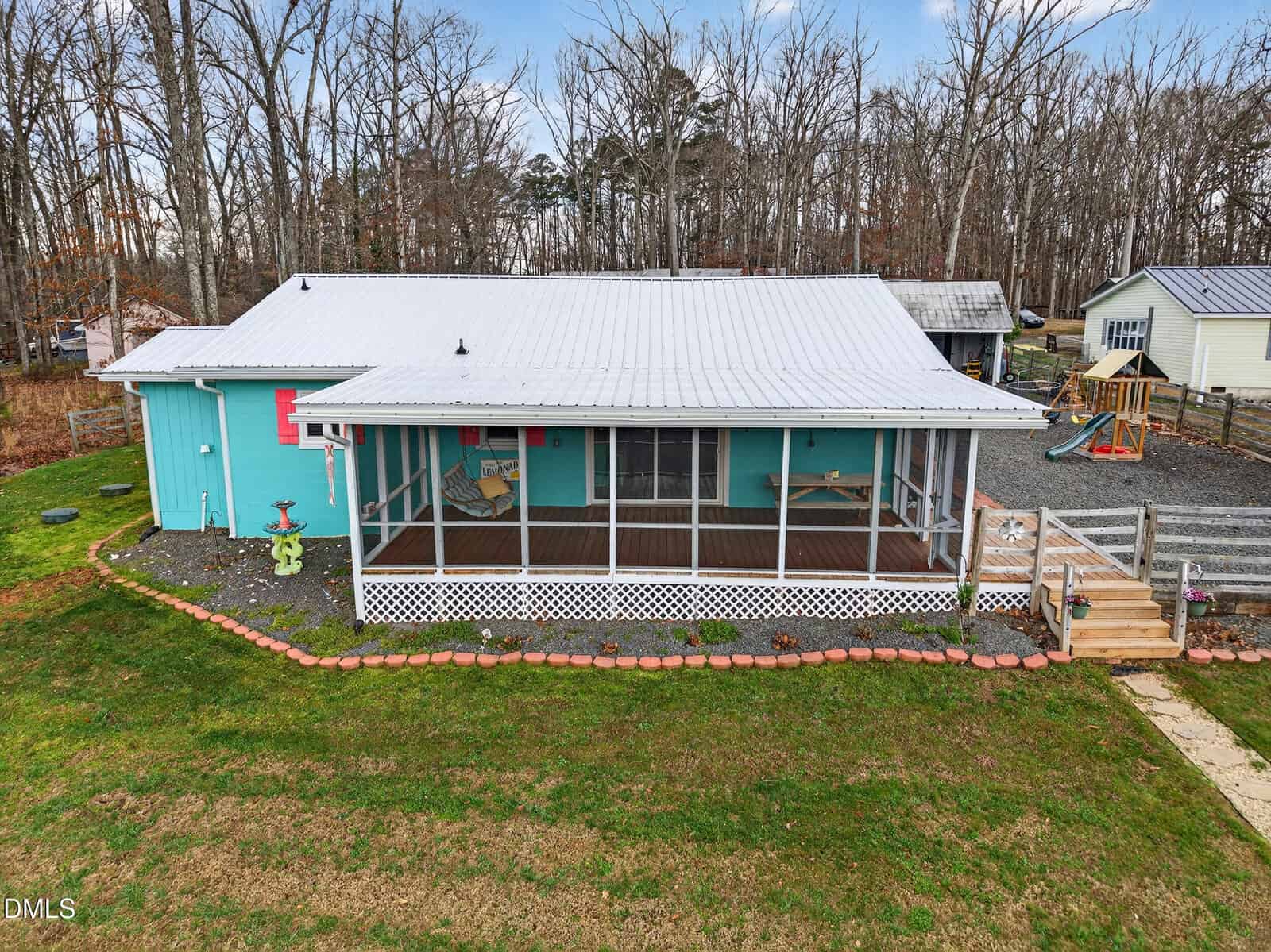 A turquoise single-story house with a white metal roof and a screened-in front porch sits on a lawn. The yard has a stone-lined garden bed, and there is a swing set and gravel driveway in the background, with trees behind the house.
