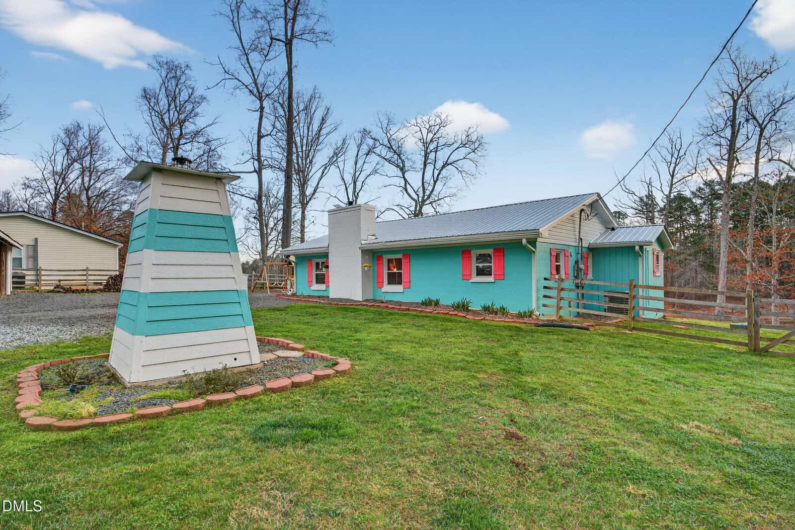 A turquoise house with red shutters and a metal roof sits on a green lawn. A white and teal well structure is in the yard. Leafless trees and a blue sky are in the background. A gravel driveway is visible on the left.