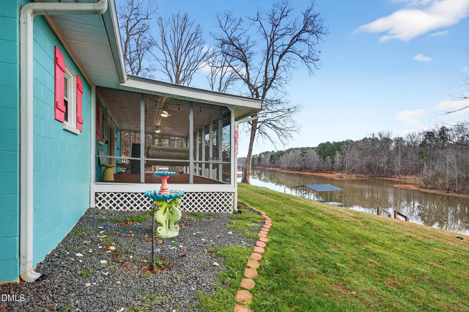 A turquoise house with red shutters and a screened porch overlooks a calm lake with a dock. There is a decorative birdbath in the yard, bordered by grass and trees, under a partly cloudy sky.