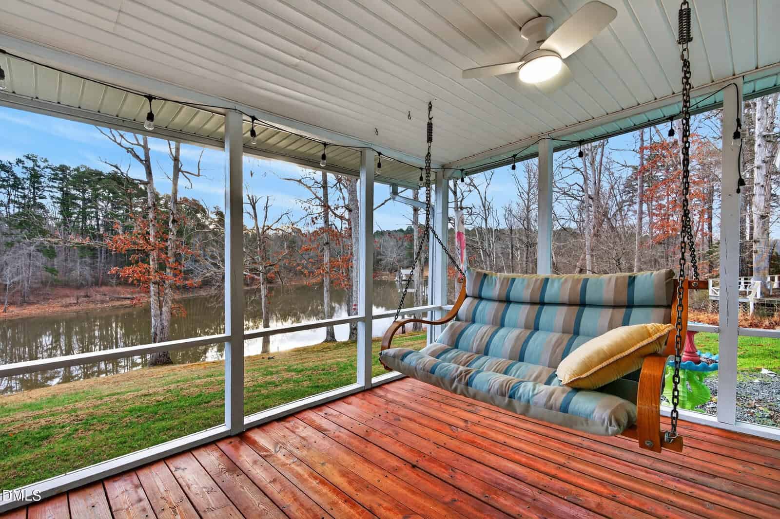 A screened-in porch with a wooden floor features a cushioned swing, overhead fan, and views of a lake and trees through the mesh panels.