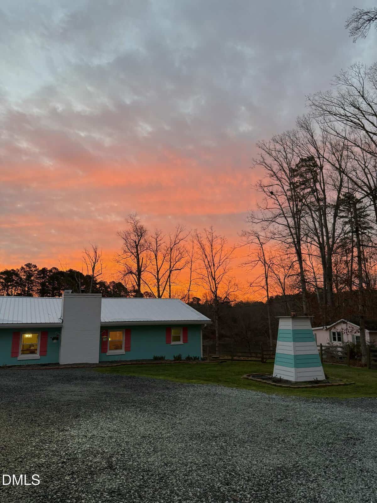 A turquoise house with white and pink trim sits under a colorful sunset sky with orange and pink clouds. Bare trees stand nearby, and a gravel driveway is in the foreground. A striped structure is to the right.