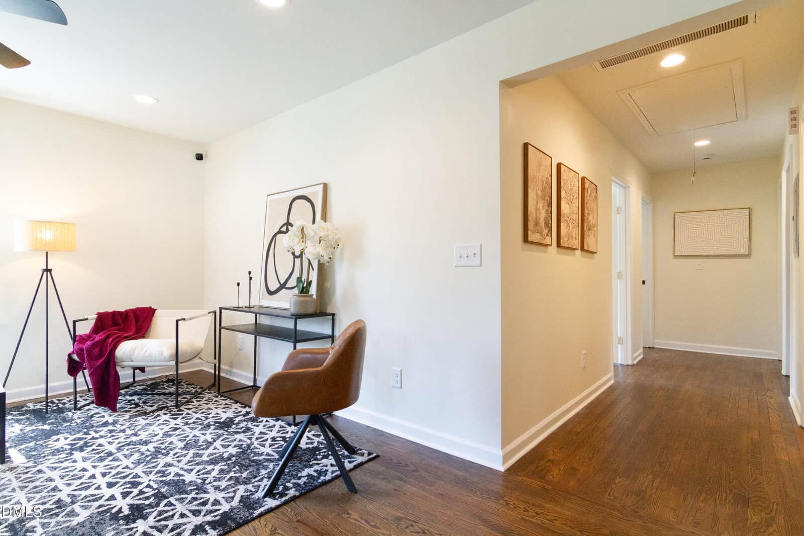 A modern home office with a desk, chair, lamp, and artwork on the wall. A hallway with framed art leads to other rooms. The floor is dark wood, and the walls are painted light beige.