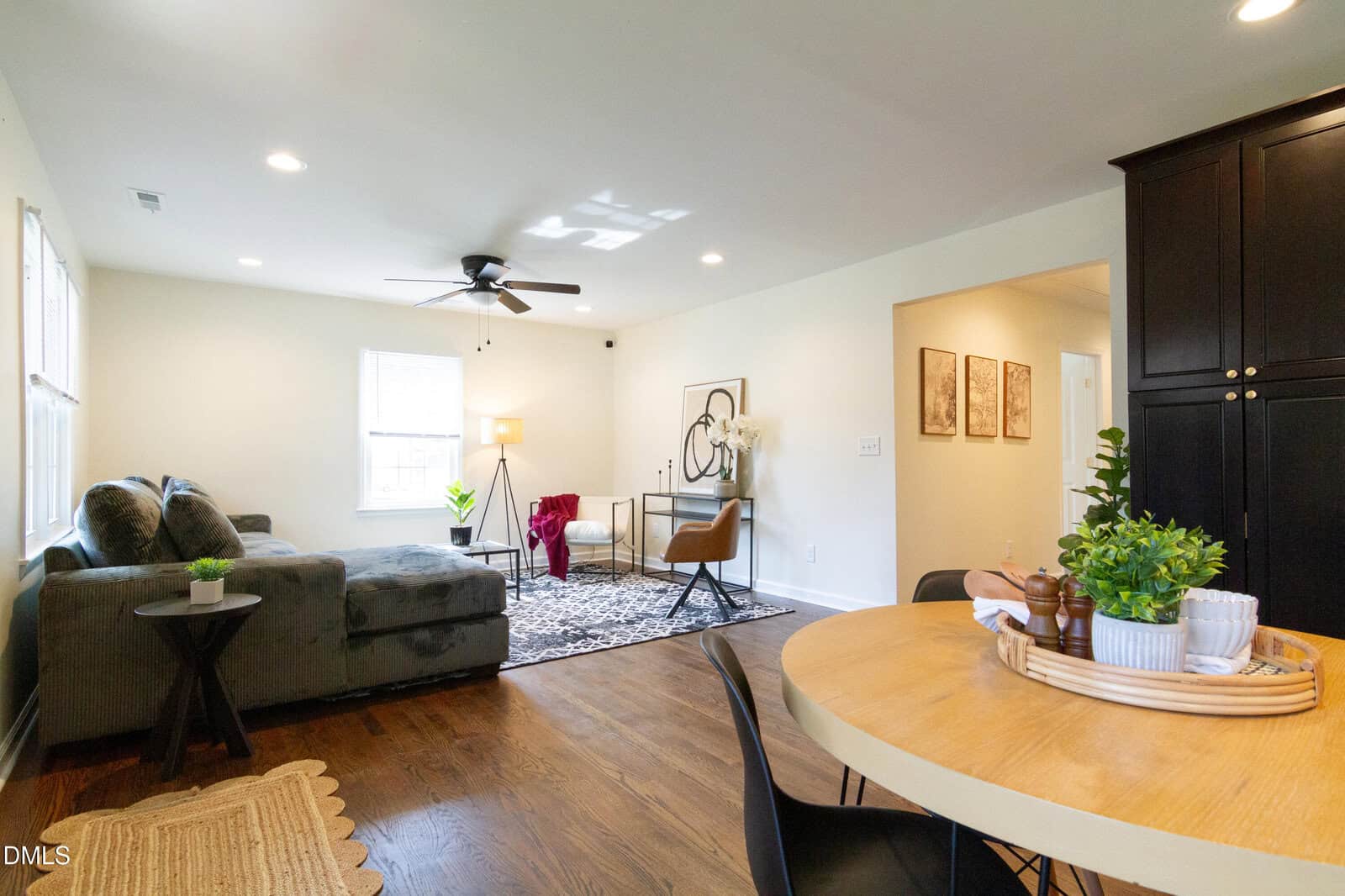 A living room with a gray sectional sofa, a patterned rug, a dark accent chair, a round dining table with plants, and wood flooring. Light walls and ceiling lights brighten the space. Art and decor add finishing touches.