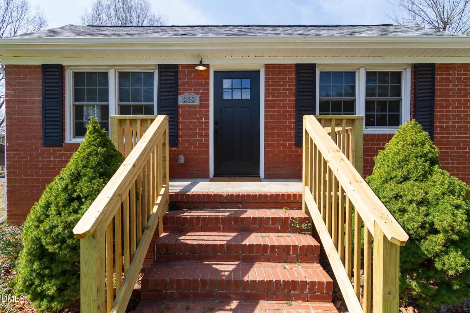A brick house on 218 Hawkins Street in Franklinton features a black front door, two windows with black shutters, a house number plaque, and a wooden railing lining brick steps. Two green shrubs flank the entrance, while trees are visible in the background.